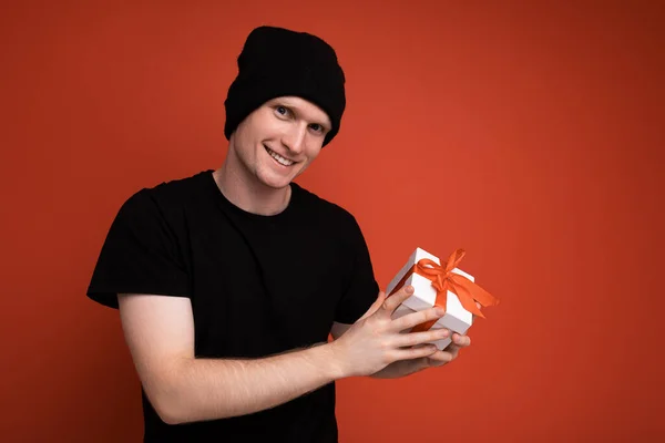 Photo shot of handsome positive smiling young male person isolated over red background wall wearing black hat and black t-shirt holding white gift box with red ribbon and looking at camera