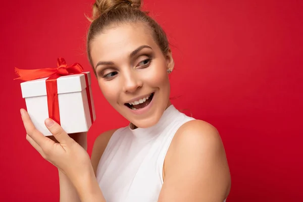 Portré fotó vonzó boldog csodálkozó fiatal szőke nő elszigetelt felett piros háttér fal visel fehér felső gazdaság ajándék doboz és keres az oldalán — Stock Fotó