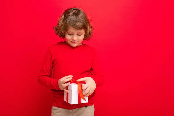 Feliz rubio rizado chico aislado sobre fondo rojo pared usando suéter rojo celebración de caja de regalo y mirando presente caja — Foto de Stock