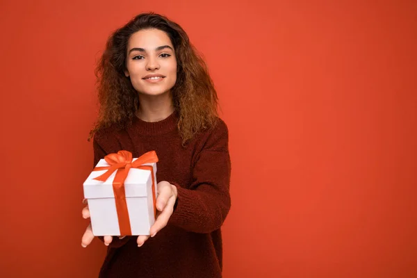 Gyönyörű boldog fiatal barna nő elszigetelt át színes háttér fal visel elegáns alkalmi ruhák gazdaság ajándék doboz és nézi a kamera — Stock Fotó