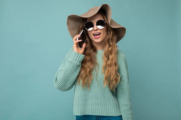 Foto de la encantadora mujer rizada rubia positiva joven con suéter azul sombrero y gafas de sol de pie aislado sobre fondo azul hablando por teléfono —  Fotos de Stock