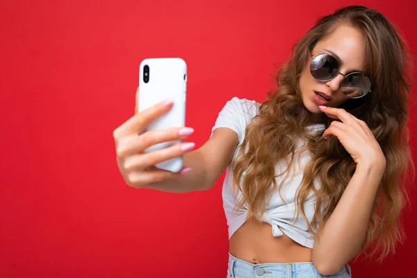 Foto de primer plano de la increíble hermosa mujer rubia joven sosteniendo el teléfono móvil tomando foto selfie utilizando la cámara del teléfono inteligente con gafas de sol traje elegante todos los días aislado sobre fondo de pared colorido — Foto de Stock