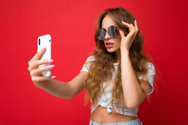 Incredibile bella giovane donna in possesso di telefono cellulare scattare foto selfie utilizzando la fotocamera dello smartphone indossando occhiali da sole ogni giorno elegante vestito isolato su sfondo parete colorato guardando dispositivo — Foto Stock