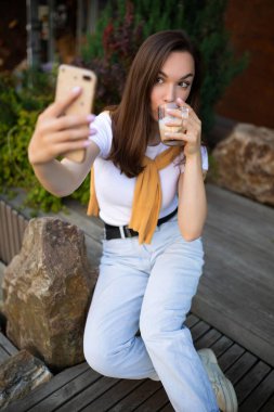 Genç kadın serbest çalışan parkta oturuyor ve elinde telefonla kahve molasından zevk alıyor.