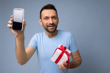 Yakışıklı, yakışıklı, yakışıklı, şık kıyafetler giyen, renkli arka plan duvarında tek başına duran, akıllı telefon tutan ve boş ekran ekranlı telefonu gösteren yakışıklı bir adam.