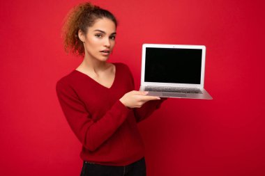 Yan profil fotoğrafı. Çekici, güzel, büyüleyici, kıvırcık, esmer, kırmızı süveter giyen, kırmızı duvarın arkasında duran, elinde boş bir kopya olan bilgisayar dizüstü bilgisayarını tutan bir kadın.