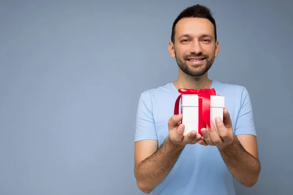 Foto de guapo positivo sonriente morena sin afeitar joven aislado sobre la pared de fondo azul con camiseta azul sosteniendo caja de regalo blanca con cinta roja y mirando a la cámara —  Fotos de Stock