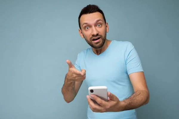 Foto de asombrado preguntando joven guapo con barba usando todos los días camiseta azul aislado sobre fondo azul sosteniendo y utilizando la comunicación del teléfono móvil en línea en Internet mirando a la cámara —  Fotos de Stock