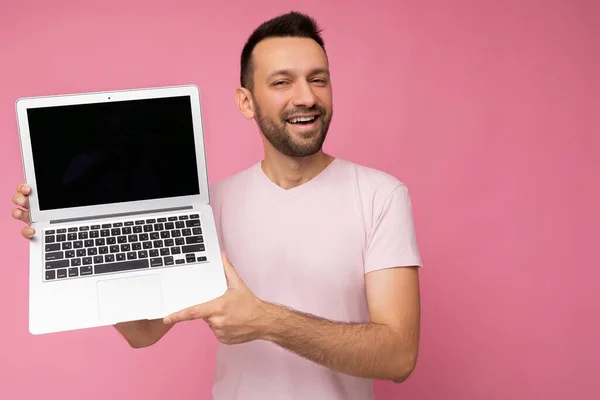 분홍색 배경에 티셔츠를 입고 컴퓨터를 들고 카메라를 보고 있는 손에 미소짓고 있는 남자 — 스톡 사진