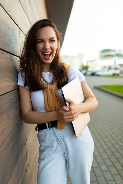 Krásný šťastný brunet mladá žena hledá na straně v ulici s notebookem v rukou s bílým tričkem a modré džíny — Stock fotografie