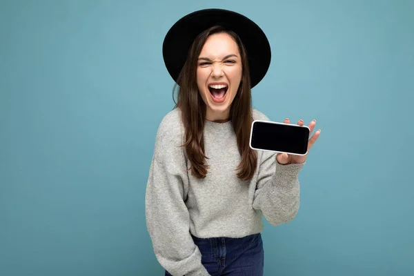 검은 모자와 회색 스웨터를 입고 있는 아름다운 젊은 여성의 초상화 사진 배경에서 고립 된 스마트폰을 보여 주는 스마트폰을 들고 있는 사진 — 스톡 사진