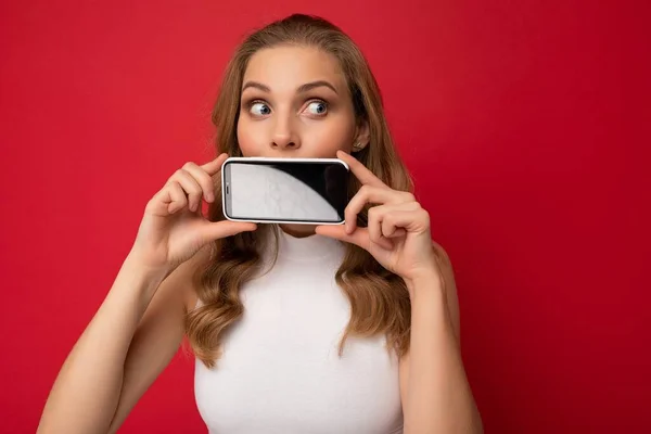 Attractive young blonde female person wearing white t-shirt isolated on red background with copy space holding smartphone showing phone in hand with empty screen for cutout looking to the side — Stock Photo, Image