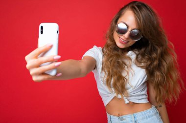 Çekici, genç, gülümseyen, mutlu bir kadın cep telefonu tutuyor ve kullanıyor. Selfie çekiyor. Duvar arkasından izole edilmiş şık kıyafetler giyiyor.