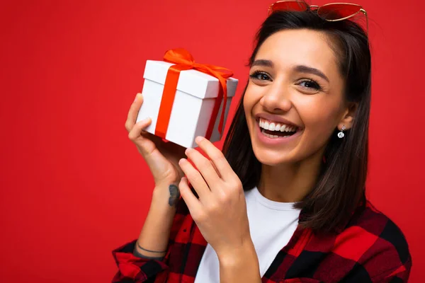 Skott av attraktiva positiva leende ung brunett kvinna isolerad över färgglada bakgrund vägg bär vardagliga trendiga outfit innehav presentförpackning och tittar på kameran — Stockfoto