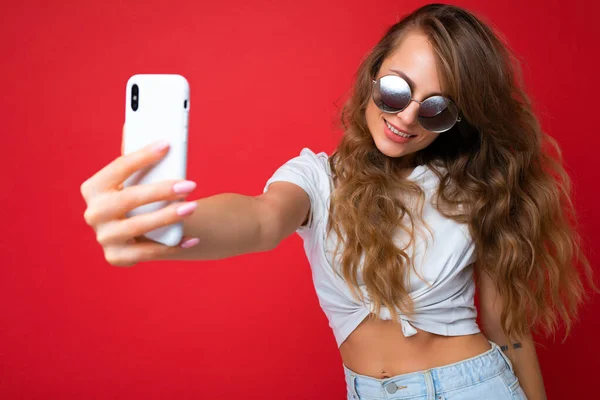Attractive charming young smiling happy woman holding and using mobile phone taking selfie wearing stylish clothes isolated over wall background — Stock Photo, Image