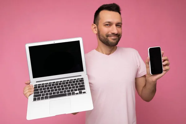 랩탑 컴퓨터와 휴대 전화를 들고 고립 된 핑크 색 배경으로 티셔츠를 입은 카메라를 어슬렁거리며 바라보고 있는 잘생긴 남자 — 스톡 사진