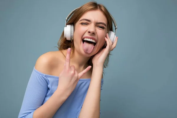 Foto de atraente jovem loira emocional vestindo top de cultura azul isolado no fundo azul vestindo fones de ouvido sem fio Bluetooth branco ouvindo música legal e se divertindo mostrando língua e — Fotografia de Stock