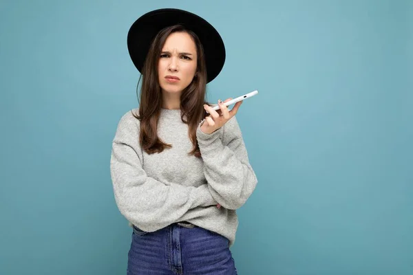 Beautiful young brunette woman thinking wearing black hat and grey sweater holding smartphone looking to the side texting isolated on background — Stock Photo, Image