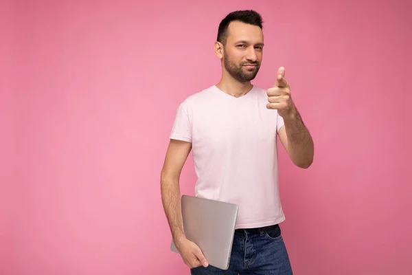 컴퓨터를 들고 핑크 색 배경으로 티셔츠를 입고 카메라를 보고 있는 모습을 가르켜 주는 잘생긴 브루 넷 남자 — 스톡 사진