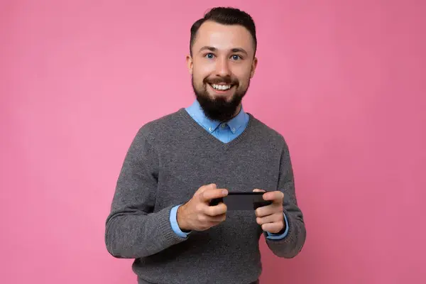 Щасливий усміхнений красивий красивий брюнетка бородатий молодий чоловік у сірому светрі та синій сорочці ізольований на рожевому фоні з порожнім простором, що тримається в руці та використовує мобільний телефонний зв'язок — стокове фото