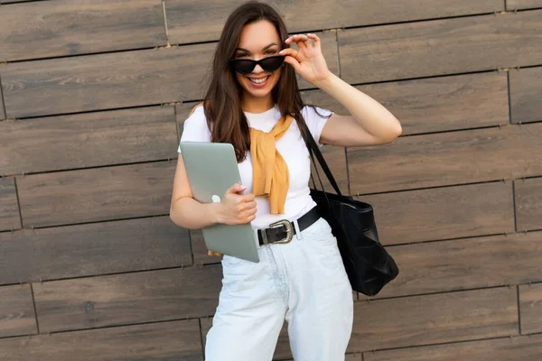 Photo of beautiful fascinating charming smiling young brunette girl wearing white t-shirt light blue jeans and yellow sweater holding computer laptop and black sunglasses staying at the street and — Stock Photo, Image