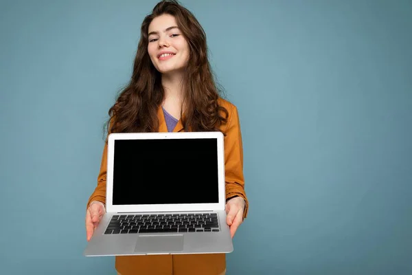 Foto de Hermosa joven feliz satisfecha sosteniendo computadora portátil mirando a la cámara con chaqueta amarilla aislada sobre fondo de pared azul —  Fotos de Stock