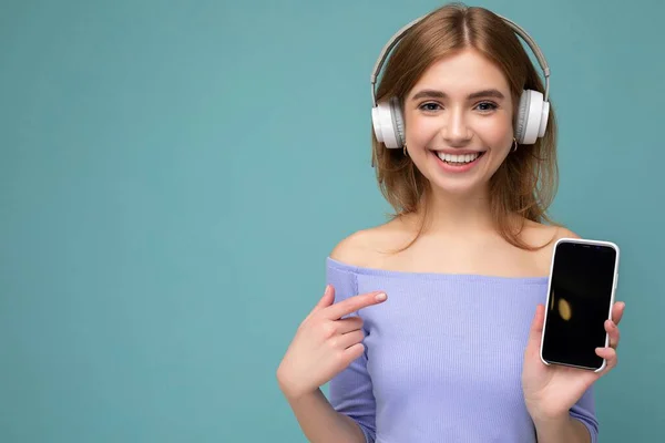 Attractive happy young blond woman wearing blue crop top isolated over blue background wall holding and showing mobile phone with empty display for mockup and wearing bluetooth headphones looking at — Stock Photo, Image