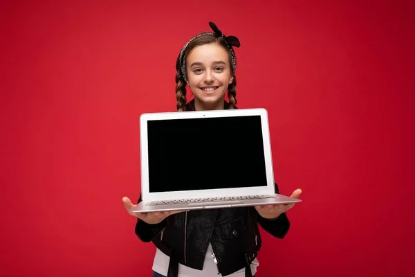 Foto de hermosa chica sonriente feliz con coletas morenas sosteniendo computadora portátil con chaqueta negra y bandana aislada sobre fondo de pared roja mirando a la cámara —  Fotos de Stock