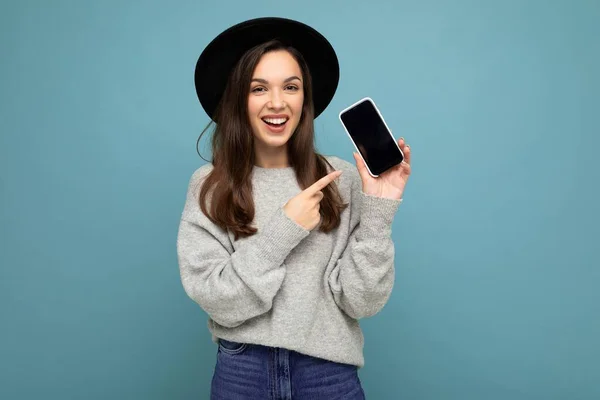 검은 모자와 회색 스웨터를 입은 매력적 인 젊은 행복 한 여성의 모습 배경 화면에서 카메라를 가리키는 손가락을 보고 있는 전화를 들고 있는 모습. — 스톡 사진