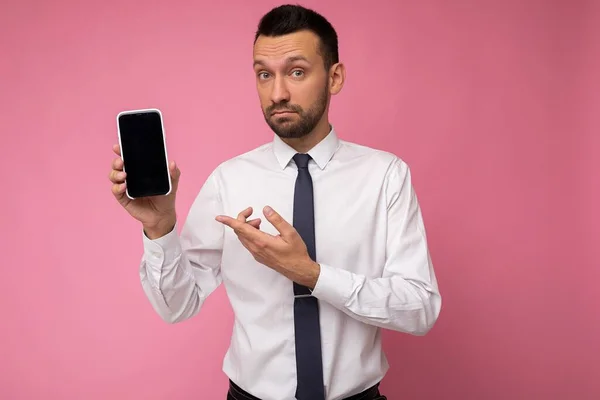 Auto-confiante bonito homem bonito vestindo camisa branca casual e gravata isolada no fundo rosa com espaço vazio segurando na mão e mostrando telefone celular com tela vazia para mockup olhando — Fotografia de Stock