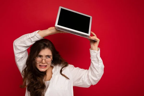 Foto de divertida hermosa cabellera oscura enojada joven mujer sosteniendo computadora portátil con pantalla de monitor vacía con maqueta y espacio de copia con camisa blanca mirando hacia abajo al lado aislado sobre rojo — Foto de Stock