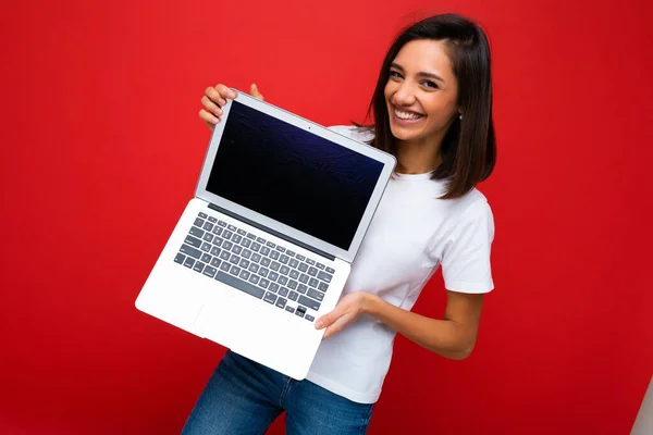 Όμορφη χαρούμενη νεαρή γυναίκα με κοντά σκούρα καστανά μαλλιά κρατώντας φορητό υπολογιστή κοιτάζοντας κάμερα φορώντας λευκό t-shirt και τζιν που απομονώνονται σε φόντο κόκκινο τοίχο — Φωτογραφία Αρχείου