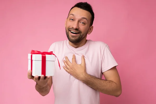 照片上，一个英俊的、积极的、面带微笑的黑发男子穿过粉红的背景墙，身穿粉色T恤，手持红丝带的白色礼品盒，看着相机 — 图库照片