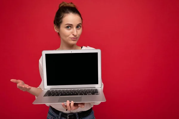 Primer plano retrato de hermosa sonriente joven feliz sosteniendo ordenador portátil mirando a la cámara que tiene preguntas con ropa elegante casual aislado sobre fondo de pared roja — Foto de Stock