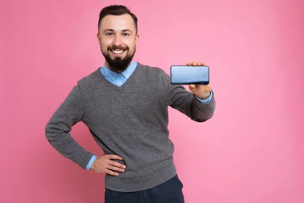 英俊而又酷的年轻黑发男子，穿着时髦的灰色毛衣和蓝色衬衫，留着智能手机，站在粉红的背景墙上，拿着手机，手里拿着手机。 — 图库照片