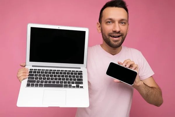 Hombre guapo con la boca abierta sosteniendo ordenador portátil y teléfono móvil mirando a la cámara en camiseta sobre fondo rosa aislado —  Fotos de Stock