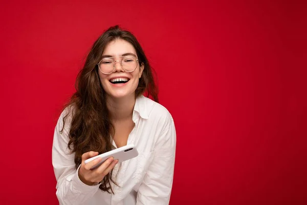 흰색 셔츠와 광학 안경을 착용하고 빨간 배경에 고립 된 예쁘게 웃고 있는 젊은 브라 운트 여성의 사진, 손에 핸드폰 게임을 하며 카메라를 보고 있는 모습 — 스톡 사진