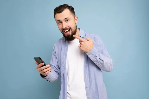 Позитивный эмоциональный красивый молодой брюнет бородатый мужчина в повседневной синей рубашке и белой футболке позирует изолирован на синем фоне с пустое пространство держит в руке мобильный телефон глядя на камеру — стоковое фото