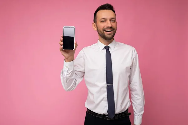 잘 생긴 잘생긴 남자가 흰색 셔츠를 입고 핑크 색 배경에 고립되어 빈 공간을 손에 들고 휴대폰을 보여 주며 흉내를 내기 위해 빈 화면을 보여 주는 사진. — 스톡 사진