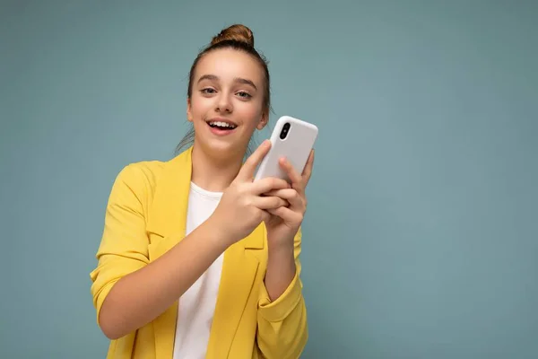 Hermosa joven sorprendida chica con chaqueta amarilla y camiseta blanca de pie aislado sobre fondo azul navegar en Internet a través del teléfono mirando a la cámara — Foto de Stock