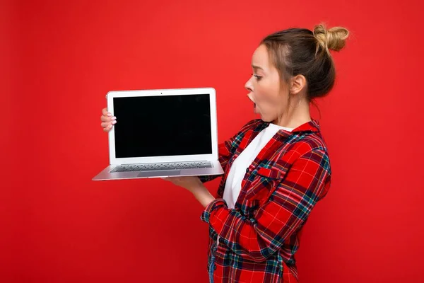 Fotografie profil lateral a Frumos șocat uimit tânără femeie care deține laptop purtând cămașă roșie uitându-se la monitor ecran izolat pe fundal roșu — Fotografie, imagine de stoc