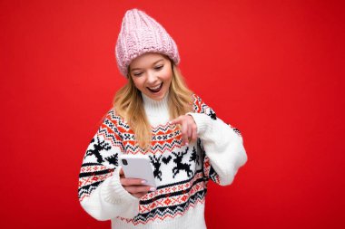 Güzel, gülümseyen sarışın bir kadının, kalın örgü şapkalı ve kalın kazaklı fotoğrafı. Kırmızı arka planda izole bir şekilde duruyor. Akıllı telefon aracılığıyla iletişim kuruyor.