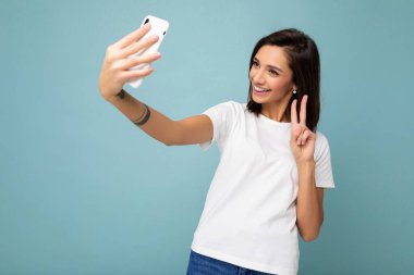 Güzel, gülümseyen, mutlu, esmer bir kadının duvar mavisi arka planda izole edilmiş bir tişört giydiği ve cep telefonuyla cep telefonu çektiği bir fotoğraf. Gadjet ekranına bakıp...