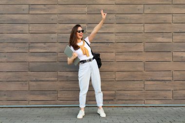 Genç, alımlı, güzel, esmer bir kadının beyaz tişörtlü ve açık mavi kot pantolonlu beyaz spor ayakkabılarıyla barış jesti yaparken çekilmiş uzun bir fotoğrafı.
