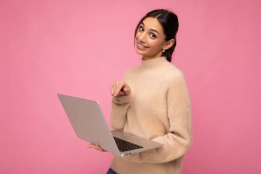Güzel, esmer bir kadının elinde bej kazak, elinde netbook bilgisayarla pembe duvar arkasında dizüstü bilgisayara bakarken çekilmiş fotoğrafı.