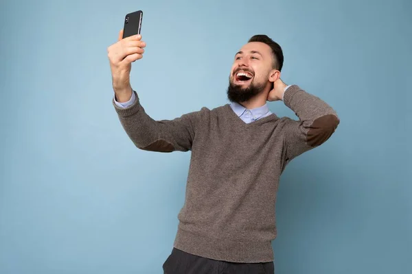 Photo of positive handsome young brunette unshaven man with beard wearing casual grey sweater and blue shirt isolated on pink background wall holding smartphone taking selfie photo looking at mobile — Stock Photo, Image