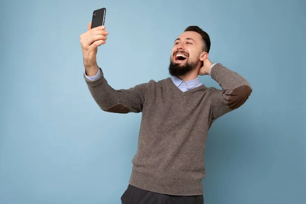 Foto de joven morena guapo positivo sin afeitar hombre con barba usando suéter gris casual y camisa azul aislado en la pared de fondo rosa sosteniendo teléfono inteligente tomando foto selfie mirando móvil —  Fotos de Stock