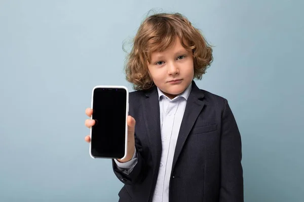 Retrato de close-up de menino bonito positivo com cabelo encaracolado usando terno segurando telefone isolado sobre fundo azul olhando para a câmera e mostrando smartphone com tela de exibição vazia — Fotografia de Stock