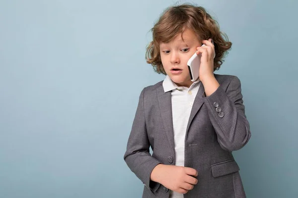 Foto van Knappe jongen met krullend haar in grijs pak vasthouden en met behulp van telefoon geïsoleerd over blauwe achtergrond naar beneden kijken en praten op mobiel met iemand — Stockfoto