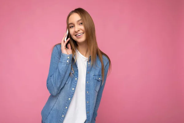 Foto van aantrekkelijke sexy positieve jonge blonde vrouw dragen casual blauw jean shirt geïsoleerd over roze achtergrond houden in de hand en praten op mobiele telefoon kijken naar camera — Stockfoto